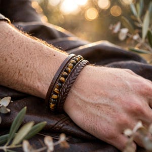 May include: A multi-strand bracelet featuring a row of round, brown and gold tiger eye beads, a smooth dark brown leather band, and a braided dark brown leather cord, all secured together as a single layered wrist accessory.