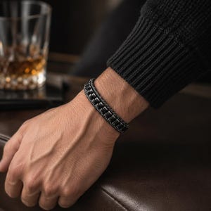 May include: A dark metallic bracelet featuring a series of interconnected, rectangular, textured links. The bracelet has a matte, gunmetal-toned finish and a structured, segmented design that wraps around the wrist, showcasing a uniform pattern.