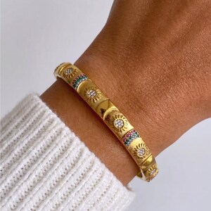 May include: A gold-toned bangle bracelet featuring a repeating pattern of embossed sun motifs inlaid with clear crystals and rectangular segments decorated with small, multi-colored stones arranged in horizontal rows along the band.