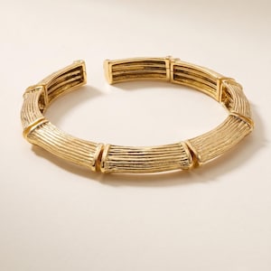 Op de afbeelding: Een goudkleurige armband met een gesegmenteerd ontwerp dat het uiterlijk van bamboestengels nabootst. Het metalen oppervlak heeft een getextureerde, geribbelde afwerking met een metaalglans en het sieraad is aan één kant open.