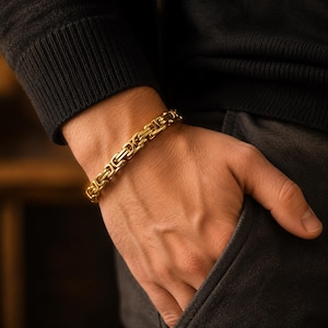 May include: A gold-toned Byzantine chain bracelet featuring a series of interlocking, square-shaped metal links. The bracelet has a polished, reflective finish and is worn around the wrist, showcasing the intricate, repeating pattern of the chain design.