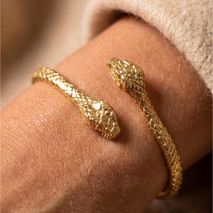May include: A gold-toned metal cuff bracelet featuring a bypass design with two detailed snake heads at each end. The surface of the bracelet is textured with a repeating scale pattern that mimics the appearance of snake skin along the entire band.