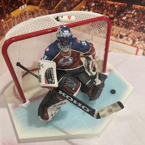 May include: A hockey goalie figurine wearing a red, white and blue uniform with a black helmet and a black and white goalie stick. The figurine is in a goalie stance with the stick in front of them. The figurine is on a white base with a blue ice rink design.