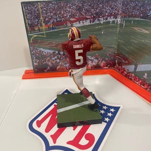 May include: A figurine of a football player in a burgundy and gold uniform, number 5, throwing a football. The figurine is on a green base with a white stripe, atop an NFL logo. A backdrop shows a football stadium.