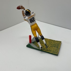 May include: A figurine of a football player in a yellow and black uniform, catching a brown football. The player is mid-air, reaching for the ball. The figurine is on a small, green, rectangular base with a red goal post.