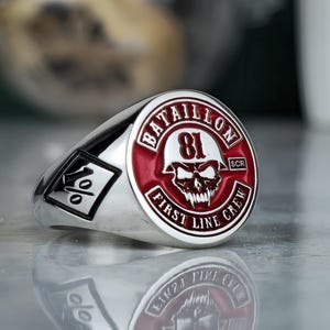 May include: A silver-toned ring featuring a circular red enamel inlay with a white skull graphic wearing a helmet, the number 81, and the text BATAILLON FIRST LINE CREW. The side of the ring displays a black and white square emblem with the number 1.