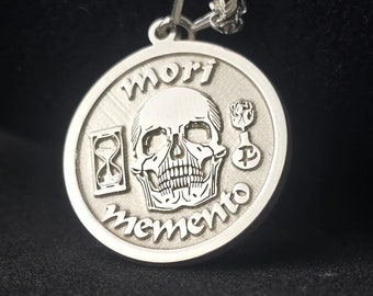 May include: Silver pendant necklace with a round charm featuring a skull, hourglass, and key. The charm is engraved with the words "Mori Memento".