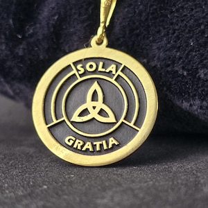 May include: A gold pendant necklace with a round, black and gold medallion. The medallion features a triquetra symbol and the words "Sola Gratia" inscribed around the edge.