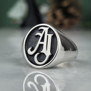 May include: A silver signet ring with a black oval face featuring the capital letters "A" and "J" in a stylized font. The ring has a polished, reflective surface and a classic design.