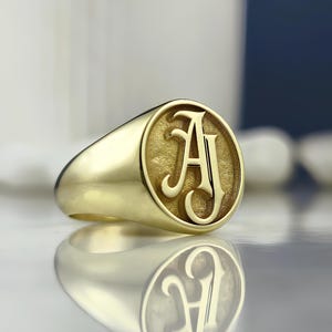 Puede incluir: Un anillo de sello dorado con la letra "A" en una elegante fuente serif. El anillo tiene una banda pulida y redondeada y una cara circular con la inicial. El anillo se muestra sobre una superficie reflectante.