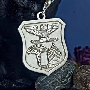 Puede incluir: Collar con colgante de plata con un dije en forma de escudo. El dije presenta un águila, un caduceo médico y una insignia de rango militar. El texto "Devil Doc 32 Years" está grabado en el dije.