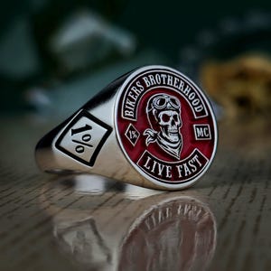 Peut inclure: Bague de motard argentée avec un motif de crâne rouge et blanc. La bague porte les inscriptions "BIKERS BROTHERHOOD" et "LIVE FAST". La bague comporte également un emblème en forme de losange avec le texte "1%" sur le côté.
