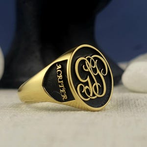 Może przedstawiać: Złoty pierścień sygnetowy z czarną owalną górą, na której znajduje się monogram liter "GG" w ozdobnym kroju pisma. Pierścień jest grawerowany słowem "ACritter" z boku.