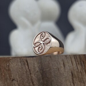 May include: A rose gold signet ring featuring a circular face with a textured background and an engraved symmetrical swirling knot design. The band is smooth and polished, showcasing a warm metallic finish that reflects light across its surface.