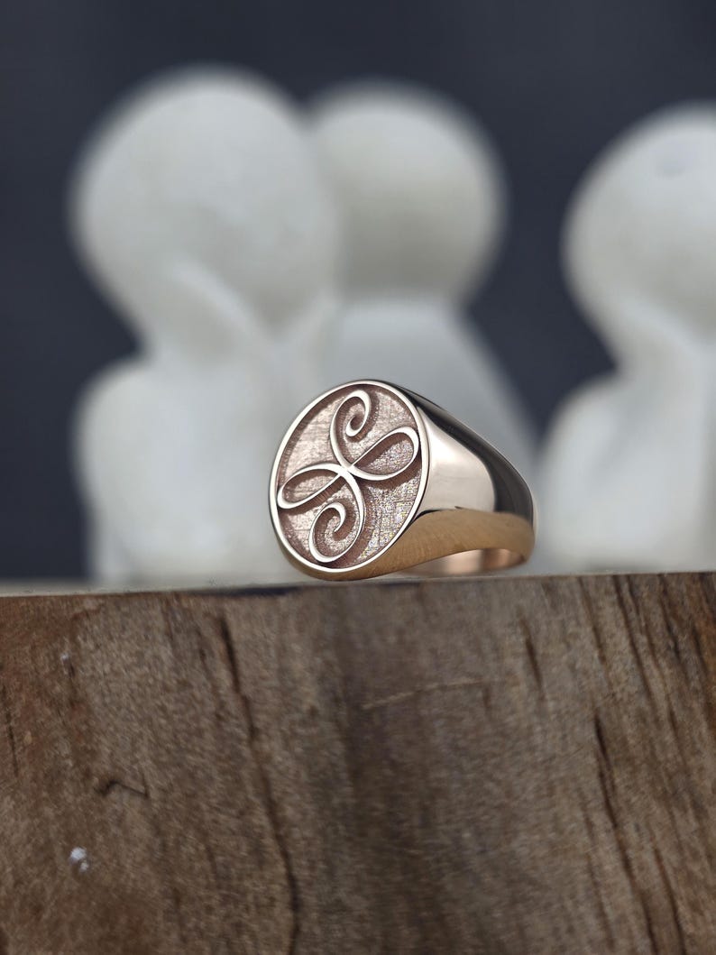 May include: A rose gold signet ring featuring a circular face with a textured background and an engraved symmetrical swirling knot design. The band is smooth and polished, showcasing a warm metallic finish that reflects light across its surface.