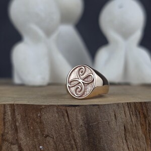 May include: A rose gold signet ring featuring a circular face with a textured background and an engraved, symmetrical, four-petaled floral or knot-like design centered on the surface. The band is smooth and polished with a warm metallic finish.