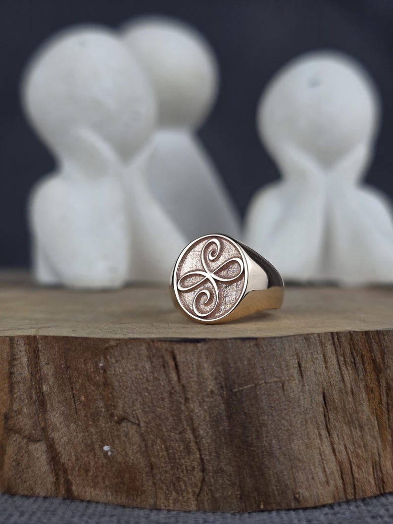 May include: A rose gold signet ring featuring a circular face with a textured background and an engraved, symmetrical, four-petaled floral or knot-like design centered on the surface. The band is smooth and polished with a warm metallic finish.