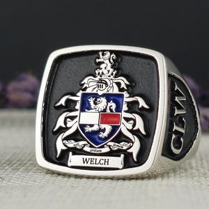 Może przedstawiać: Srebrny pierścień z czarną emaliowaną wkładką przedstawiającą herb z lwem, tarczą i napisem "WELCH" poniżej. Pierścień ma napis "CLAN" z boku.