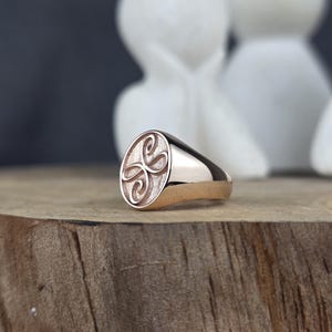 A high-detail close-up of a solid 10k pink gold signet ring with a Zibu friendship symbol. The oval face displays a stylized loop design with a dual-texture finish: a matte, grainy background with high-gloss raised detailing. The ring is set on a wooden board with a soft bokeh of white decorative figures behind it.