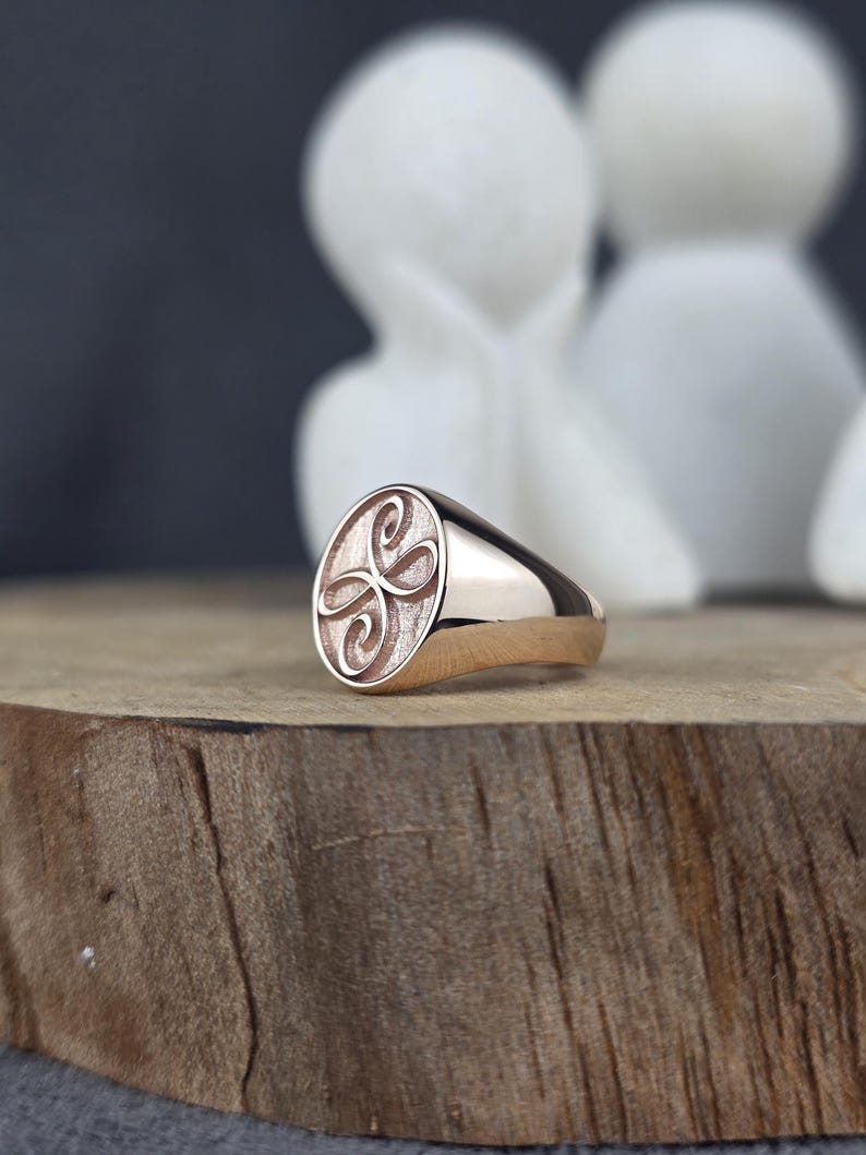 A high-detail close-up of a solid 10k pink gold signet ring with a Zibu friendship symbol. The oval face displays a stylized loop design with a dual-texture finish: a matte, grainy background with high-gloss raised detailing. The ring is set on a wooden board with a soft bokeh of white decorative figures behind it.