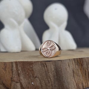 May include: A rose gold signet ring featuring a circular face with a textured background and an engraved infinity-style knot symbol. The ring has a polished metal band and a smooth, rounded silhouette, showcasing a metallic finish throughout.