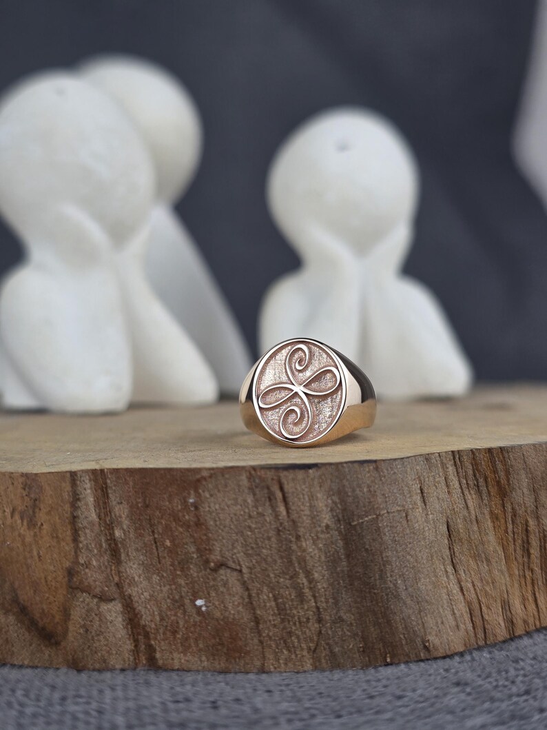May include: A rose gold signet ring featuring a circular face with a textured background and an engraved infinity-style knot symbol. The ring has a polished metal band and a smooth, rounded silhouette, showcasing a metallic finish throughout.