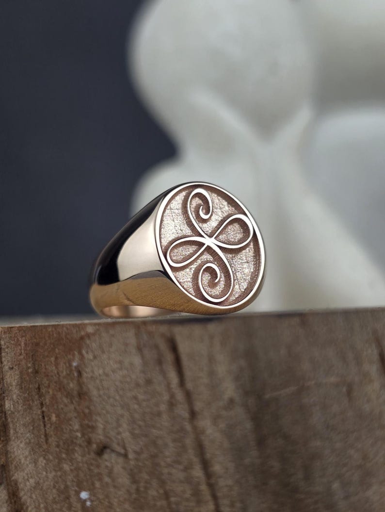 A close-up photograph of a dainty 10k rose gold signet ring featuring the engraved Zibu angelic symbol for friendship. The oval ring face displays the raised, highly polished continuous loops of the friendship symbol over a textured, matte pink gold background. The ring rests on a rustic wooden surface with a soft-focus background of white decorative ceramic figurines.