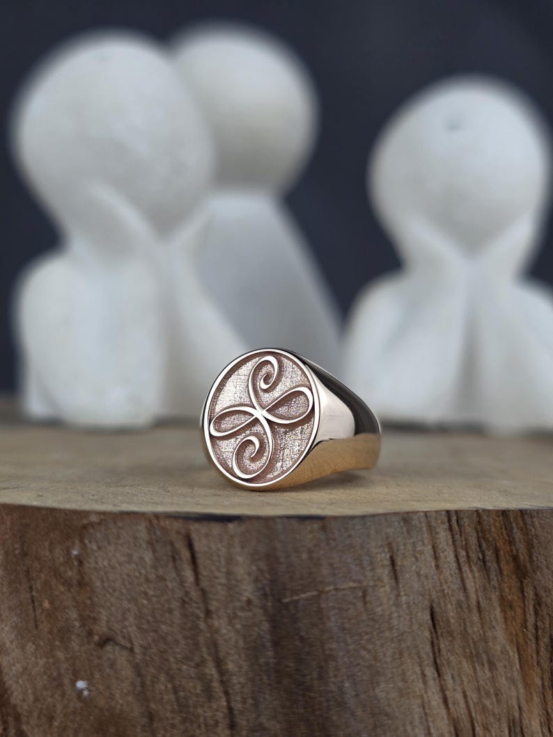 An angled close-up of a dainty 10k rose gold ring featuring a Zibu angelic symbol engraving. The oval signet face combines a textured background with a polished, raised scroll ornament. The high-polish shank of the ring reflects light cleanly as it rests on a wooden block against a dark, blurred background.