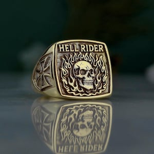 May include: A gold-colored ring with the words "HELL RIDER" above a skull engulfed in flames. The ring has a square face and a cross design on the side. The ring is highly reflective.