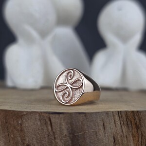 This close-up showcases a handcrafted rose gold signet ring with a Zibu symbol of friendship. The polished outer rim frames a stippled inner face where the continuous loop symbol is raised in a mirror-like finish. The ring's smooth, reflective band is visible as it stands on a wooden ledge, emphasizing its modern spiritual aesthetic.