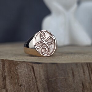 An elegant close-up of a 10k rose gold angelic symbol signet ring. The oval face is detailed with a raised Zibu friendship scroll. The contrasting textured inner surface makes the polished gold emblem stand out beautifully. The ring rests on a raw wood block, creating a warm, organic setting with blurred white decor shapes in the background.