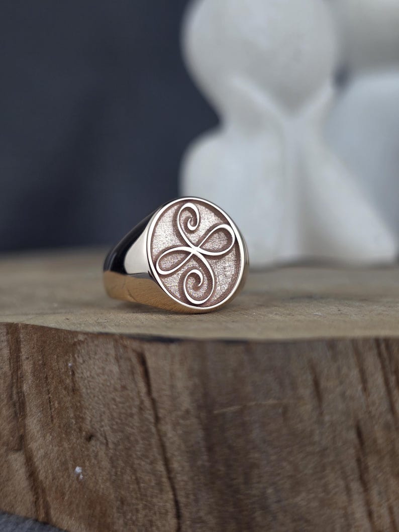 An elegant close-up of a 10k rose gold angelic symbol signet ring. The oval face is detailed with a raised Zibu friendship scroll. The contrasting textured inner surface makes the polished gold emblem stand out beautifully. The ring rests on a raw wood block, creating a warm, organic setting with blurred white decor shapes in the background.