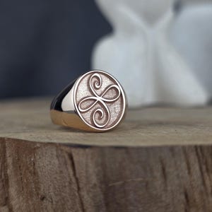 This product shot shows a minimalist 10k solid rose gold Zibu friendship ring from an angled perspective. The ring is set vertically on a natural wooden block, highlighting its polished band and bezel. The angelic symbol is raised in high relief on the oval face, contrasting against a dark, textured gold background under soft studio light.