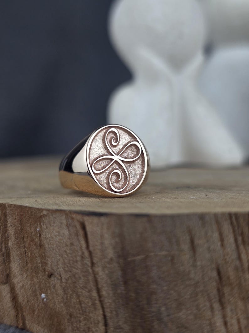 This product shot shows a minimalist 10k solid rose gold Zibu friendship ring from an angled perspective. The ring is set vertically on a natural wooden block, highlighting its polished band and bezel. The angelic symbol is raised in high relief on the oval face, contrasting against a dark, textured gold background under soft studio light.
