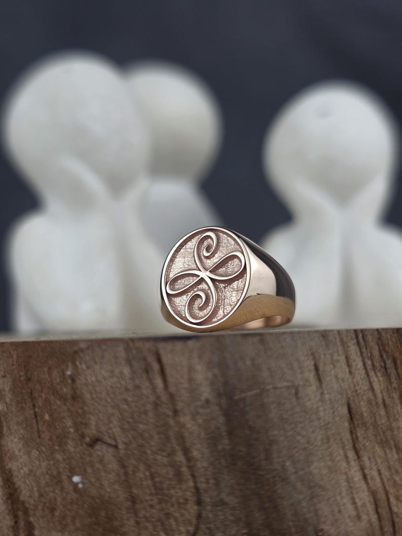 A macro shot of a minimalist rose gold Zibu angelic ring. The ring’s face features a raised four-loop friendship emblem on a finely textured, matte gold background. The outer border and polished band of the signet ring shine under soft lighting, presented as a delicate, daily spiritual accessory on a rustic wood piece.