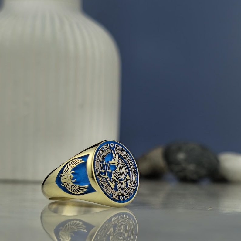 Channel the power of Anubis with this custom-crafted signet ring, where the god of the underworld is depicted in remarkable detail. Surrounded by hieroglyphs and holding scales of judgment, the design invokes justice, transformation, and ancient protection. The combination of black and gold accents set against a royal blue background creates a striking aesthetic that pays homage to millennia-old Egyptian traditions.