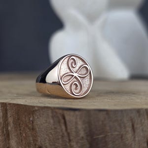 This image captures a custom rose gold Zibu friendship ring with a delicate loop design. The oval face shows a raised, continuous symbol with a polished finish, bordered by a shiny pink gold bezel. The background of the engraving has a stippled, antique texture. The ring is styled on a wooden block, showcasing its premium handcrafted details.