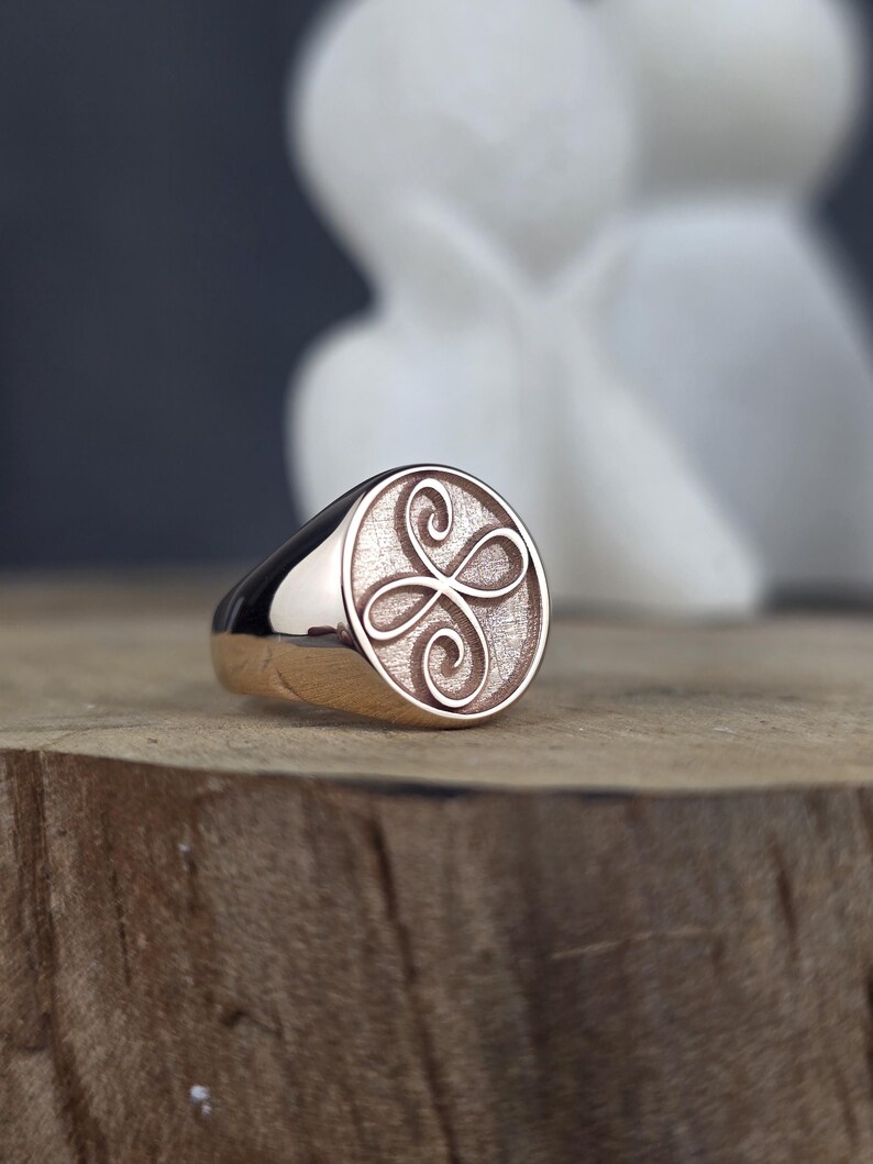 This image captures a custom rose gold Zibu friendship ring with a delicate loop design. The oval face shows a raised, continuous symbol with a polished finish, bordered by a shiny pink gold bezel. The background of the engraving has a stippled, antique texture. The ring is styled on a wooden block, showcasing its premium handcrafted details.