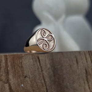 May include: A gold signet ring featuring a circular face with a textured background and an engraved four-loop infinity knot design. The band is smooth and polished, showcasing a warm metallic finish that reflects light across its curved surface.