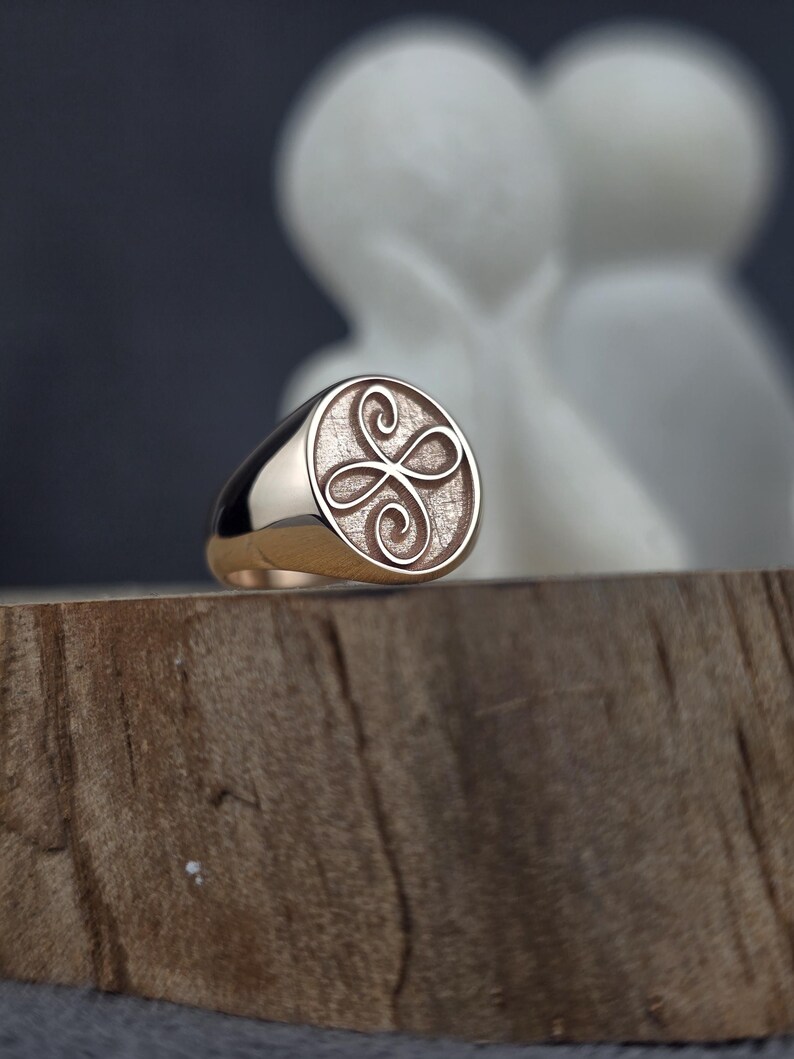 May include: A gold signet ring featuring a circular face with a textured background and an engraved four-loop infinity knot design. The band is smooth and polished, showcasing a warm metallic finish that reflects light across its curved surface.