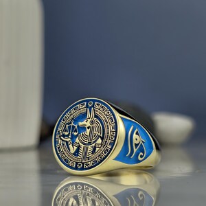 Celebrate ancient heritage with a ring that embodies the might and mystique of Anubis. Known as the guardian of the dead, Anubis is presented here with artisan clarity, featuring balanced scales and ornamental hieroglyphics that radiate from a vibrant blue background. This luxurious piece captures the essence of both ancient faith and modern fashion in one symbolic masterpiece.