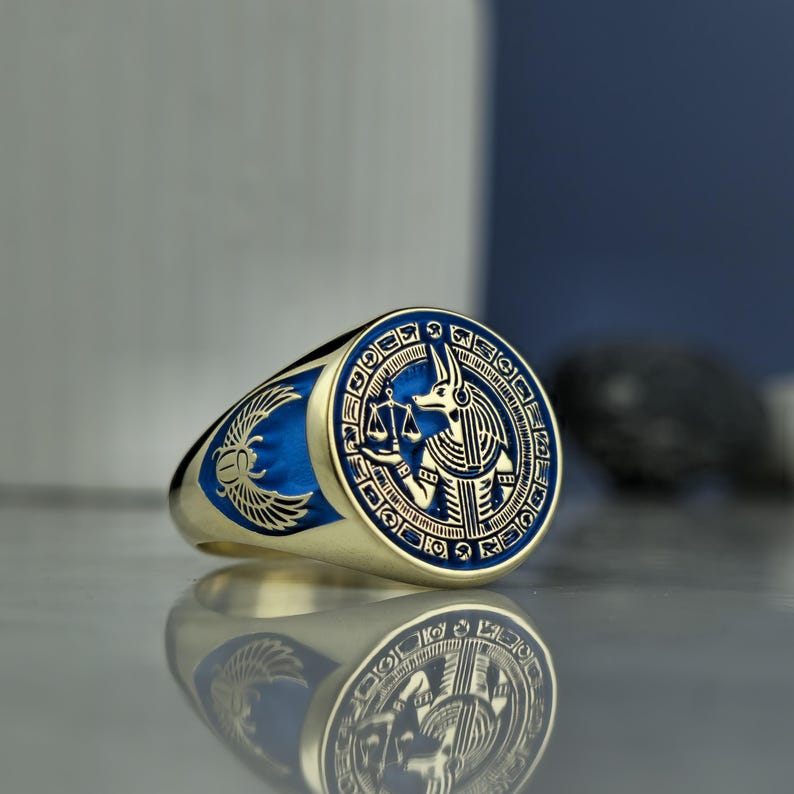 Elevate your jewelry collection with this handcrafted Anubis signet ring, inspired by the ancient Egyptian deity known for guiding souls to the afterlife. The fine gold plating and deep blue enamel add a modern touch to its mythological roots. Each detail of the design—from the weighing scale to the hieroglyphic border—celebrates Anubis’s iconic role in judgment and protection. A meaningful gift for those drawn to ancient lore, spirituality, and regal aesthetics.