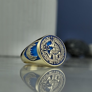 May include: A gold ring with blue enamel detailing. The ring showcases an Egyptian Anubis design on the face and a scarab beetle on the side. The ring is highly reflective, with intricate detailing.