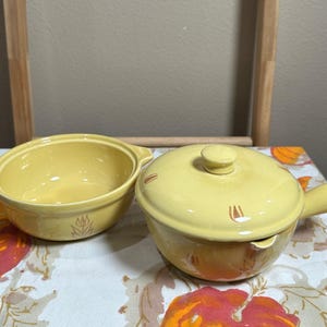 May include: A set of two yellow ceramic bowls with brown accents. The bowls have a rounded shape and a small handle on each side. The larger bowl has a lid.