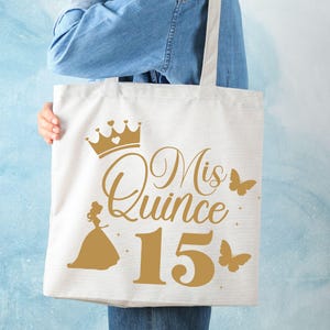 Op de afbeelding: Een witte tote bag met een gouden kroon, de tekst "Mis Quince 15" en een gouden silhouet van een meisje in een jurk. Er zijn ook gouden vlinders op de tas.
