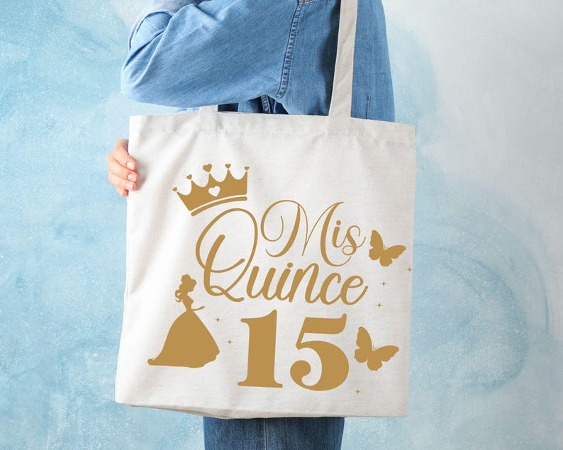 Op de afbeelding: Een witte tote bag met een gouden kroon, de tekst "Mis Quince 15" en een gouden silhouet van een meisje in een jurk. Er zijn ook gouden vlinders op de tas.