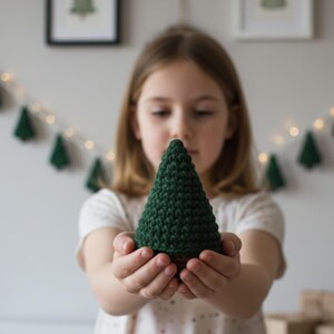 May include: A handmade, dark green crochet Christmas tree with a brown base. The tree is held in a person's hands, with a blurred background featuring Christmas decorations. The tree is approximately 5 inches tall.