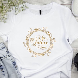 Op de afbeelding: Wit T-shirt met een gouden afbeelding van een kroon en de tekst "Mis Quince Anos" in een decoratieve cirkel.