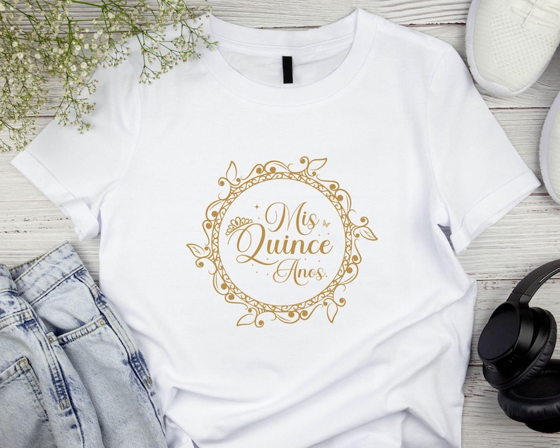 Op de afbeelding: Wit T-shirt met een gouden afbeelding van een kroon en de tekst "Mis Quince Anos" in een decoratieve cirkel.