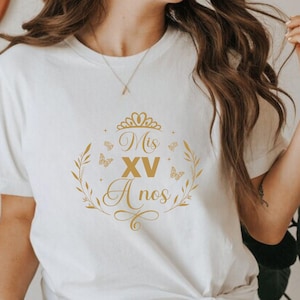 Op de afbeelding: Wit T-shirt met een gouden grafisch ontwerp met de tekst "Mis XV A&ntilde;os" met een kroon en vlinders.